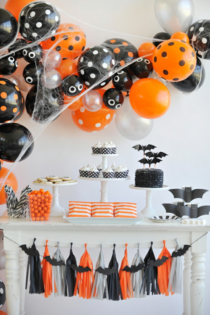 table de fête blanche, déco de halloween, gâteaux avec chauve-souris en plateaux de présentation, bonbons oranges, franges décoratives 