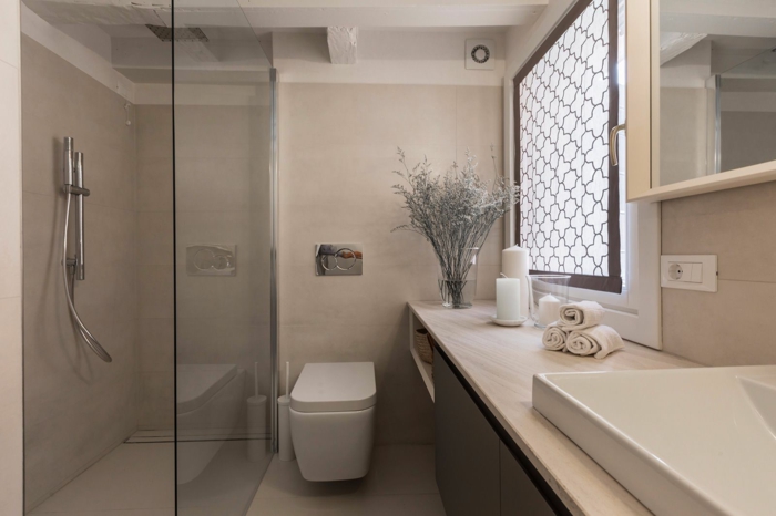 salle d'eau couleur grège, toilette blanche, plateau beige lisse, vasque rectangulaire en pente, serviettes enroulées, fenêtre avec panneau décoratif