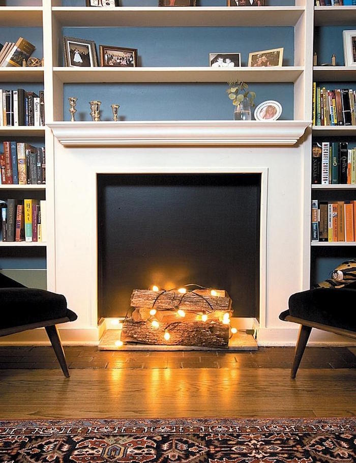meuble déco fausse cheminée avec insert fond noir et buches décoratives avec guirlande lumineuse et grande etageres bibliotheque sur mur bleu et sol parquet