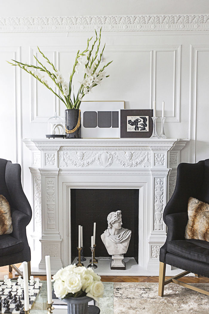 cheminée fausse avec manteau sculpté style classique dans salon blanc luxe design avec statuette buste antique et bougies dans insert à fond noir et cadre avec sol en carrelage marbre