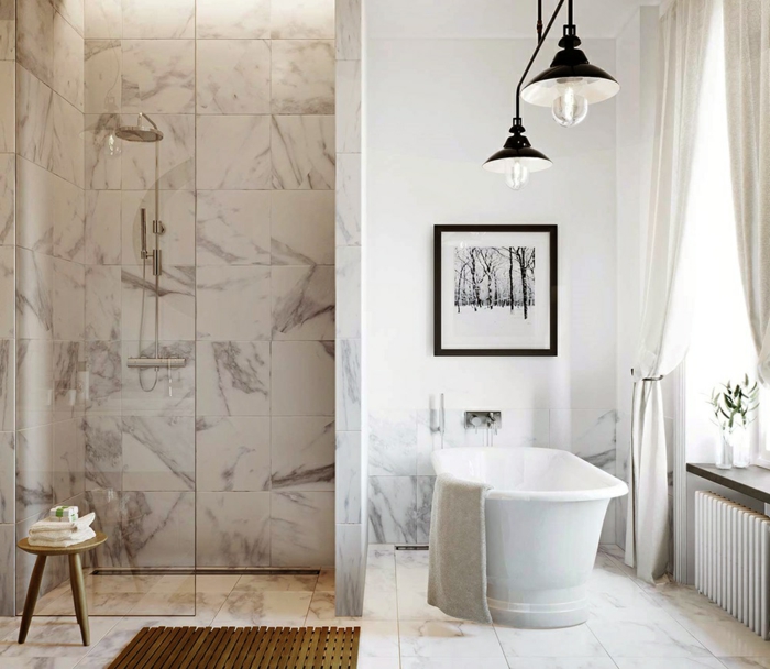 salle de bain avec une douche à l'italienne, petit tabouret en bois, baignoire blanche, lampes industrielles, cadre peinture monochrome, carreaux imitation marbre
