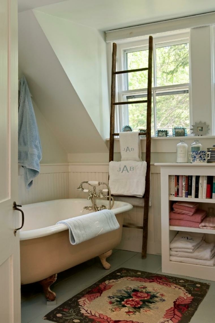 petit tapis aux motifs floraux, baignoire pastel, robinet chromé, échelle en bois, petit meuble de rangement avec livres et serviettes