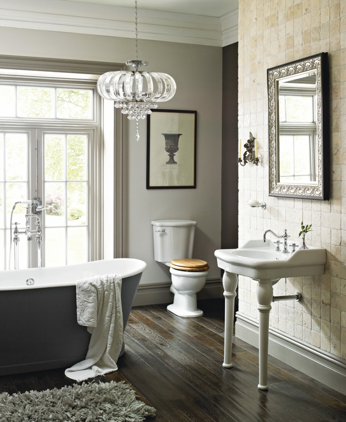 plafonnier en cristal, grande baignoire en gris et blanc, vasque vintage blanche, sol en parquet foncé, miroir rectangulaire au cadre, salle de bain imposante