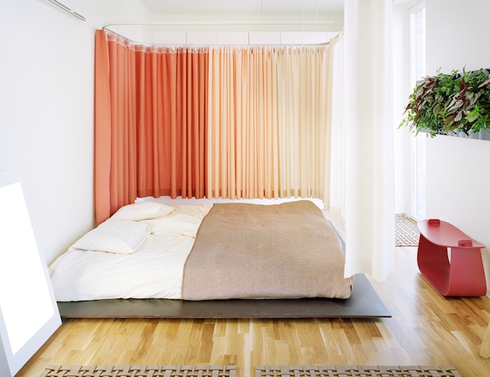 rideaux séparateurs couleur saumon et orange, lit sur plate-forme bois foncé, matelas blanc, couverture de li marron, parquet bois clair, tableau végétal