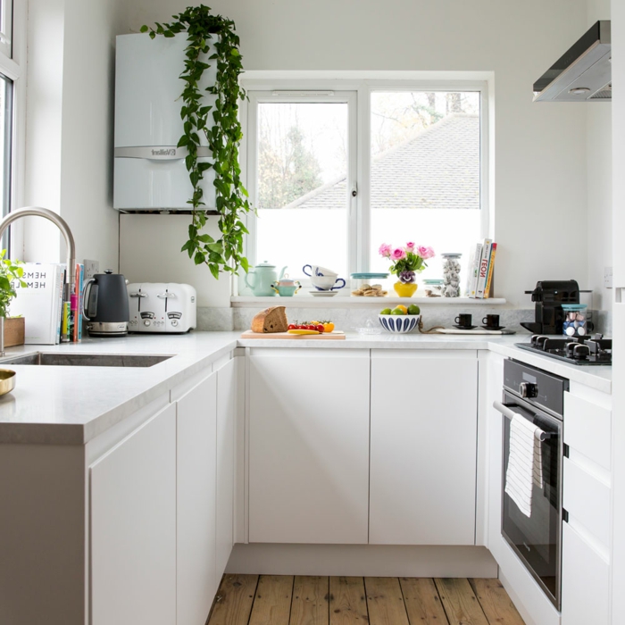 jolie cuisine blanche, sol en bois, placards blancs, évier carré, petit frigo suspendu, étagère blanche sous la fenêtre