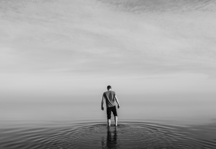 portrait noir et blanc d'un jeune homme pieds nus dans l'eau calme de la mer, en arrière plan l'horizon caché dans le brouillard