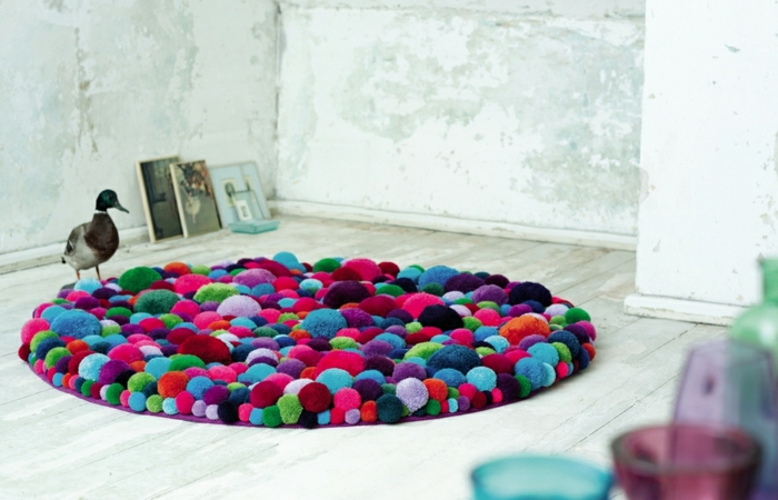pompons en laine assemblés en tapis rond multicolore, sol en lattes blanches, murs en béton, bouteilles et tasses acryliques