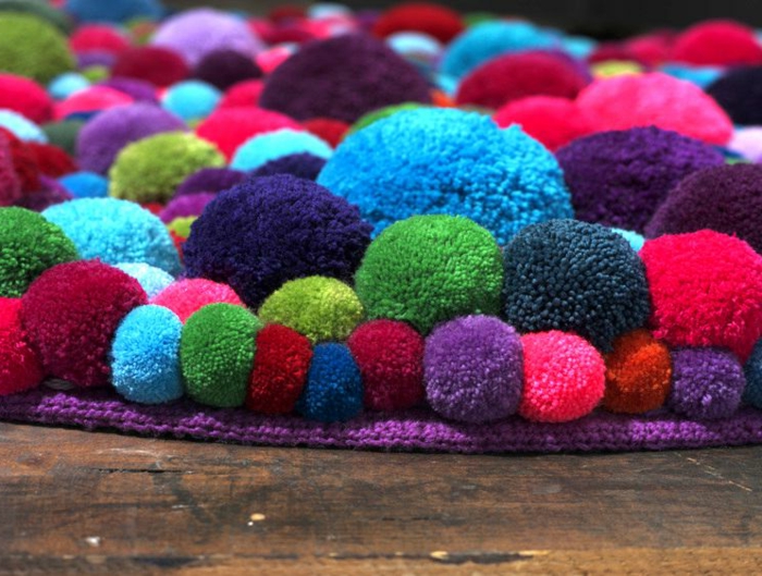 grand tapis de pompons cousus multicolores, base lilas au tricot, sol en planches, pompons de taille variable
