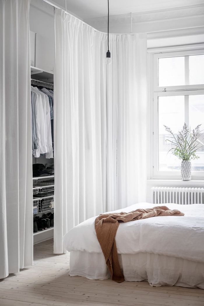 dressing avec rideau blanc qui se fond dans le décor de la chambre à coucher blanc, système de rangement pour vêtements avec barres de penderie, étagères et paniers dissimulés par des rideaux