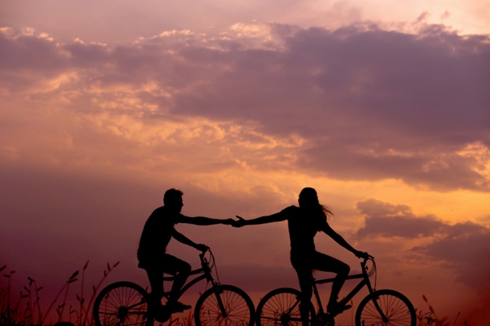 Cool photo de couple silhouettes de homme et femme qui se tiennent par la main au meme temps sur leurs bicyclettes, ciel rose-violet