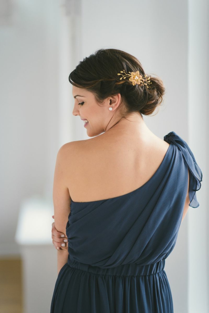robe grecque épaules asymétriques, coiffure chignon flou avec accessoire fleur, robe couleur bleu foncé