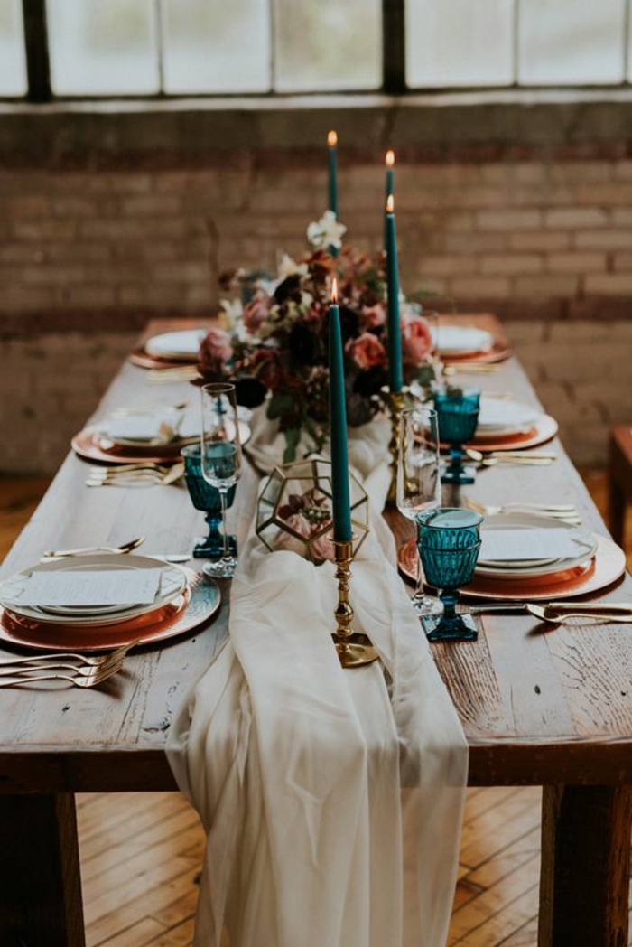 table en bois, assiettes et sous assiettes arrangées, grandes bougies turquoises, ustensiles dorés, verres bleus