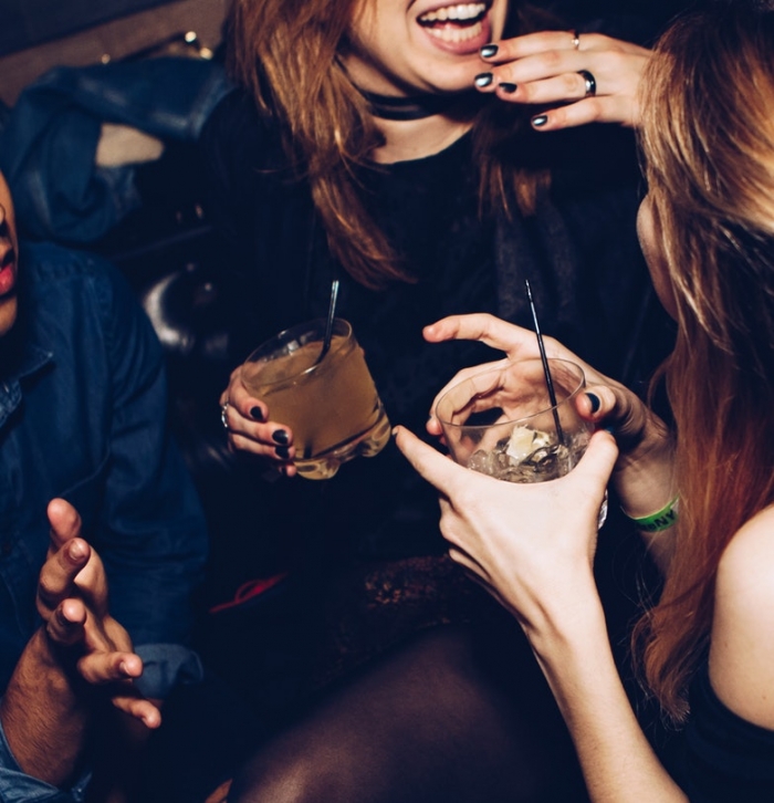 Fille souriante avec cocktail dans la main, deux filles et un homme qui parlent à une soirée