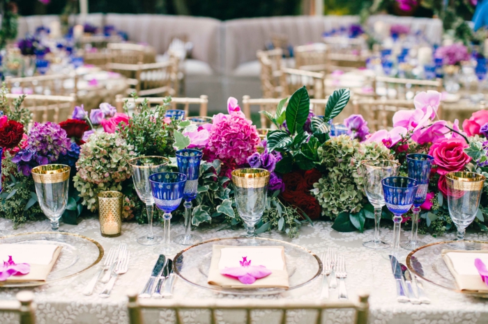 déco florale au centre de la table de mariage, verres aux rebords dorés et aux dessins bleus, assiettes transparentes, nappe blanche