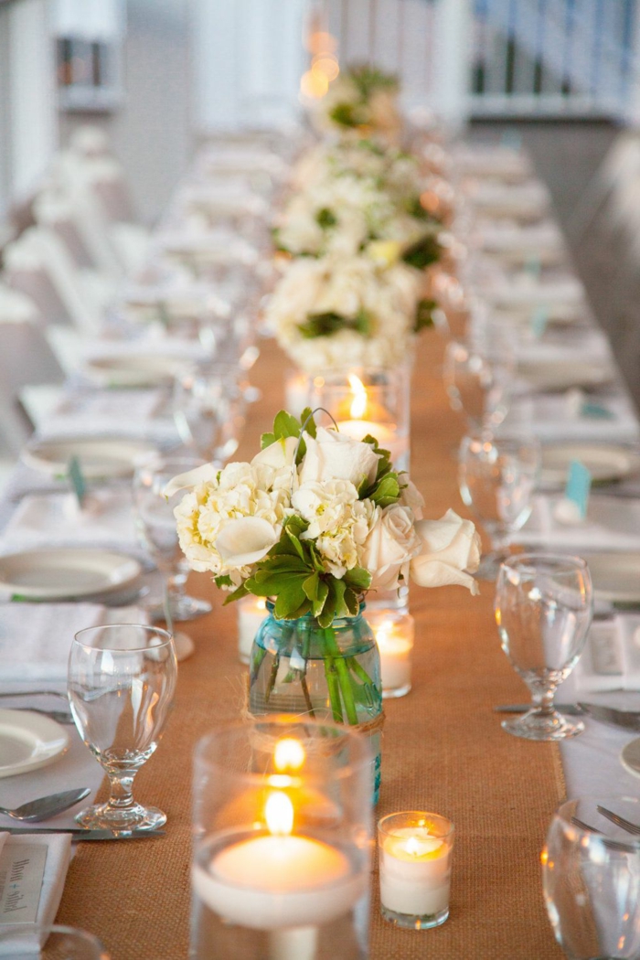 toile de jute passant par la table de mariage, bocaux de verre pleins de roses crème, verres à vin
