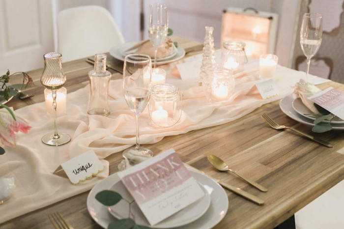 table en bois, assiettes blanches, chemin de table rose, verres élégants, chaises blanches, ustensiles dorées