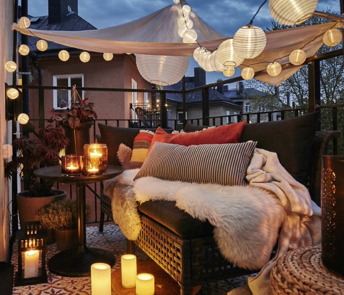 aménagement petit balcon romantique avec canapé décoré de plaid et coussins décoratifs, plusieurs bougeoirs et bougies, voile d ombrage et guirlande lumineuse boule