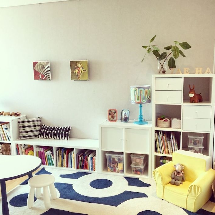 murs gris, longue etagere basse blanche pour ranger livres et boites à jouets, deco murale de cadres enfant et banc, tapis blanc et bleu marine, fauteuil jaune bas, table et tabouret bas