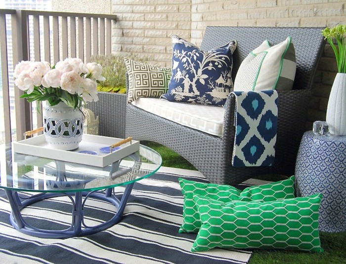 meuble balcon rotin gris, fauteul décoré de coussins blanc, bleu et vert, tapis à rayures bleu et blanc, table basse avec plateau verre, fleurs dans vase
