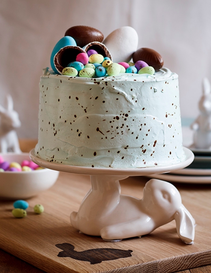 gâteau nid de pâques tacheté au glaçage de crème au beurre coloré vert pastel décoré d’œufs en chocolat, posé sur un joli présentoir à gâteau en forme de lapin