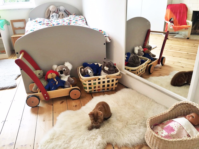 lit gris bas avec couverture colorée, miroir mural, tapis moeulleux avec chat dessus, parquet bois clair, paniers à jouets