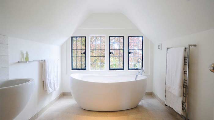 baignoire blanche dans une salle de bain en blanc avec carrelage beige clair, murs blancs, fenetres encadrement noir