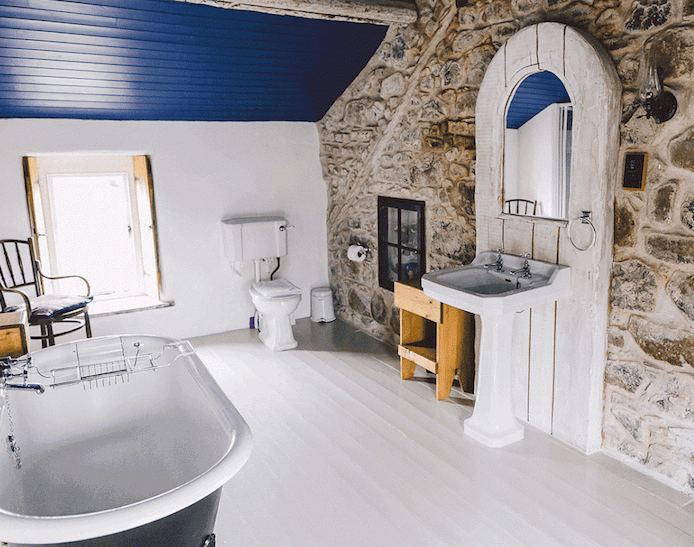 pente en lambris bleu, baignoire ancienne à poser, lavabo console blanc, sol plancher bois blanchi, mur de pierres, salle de bain sous pente