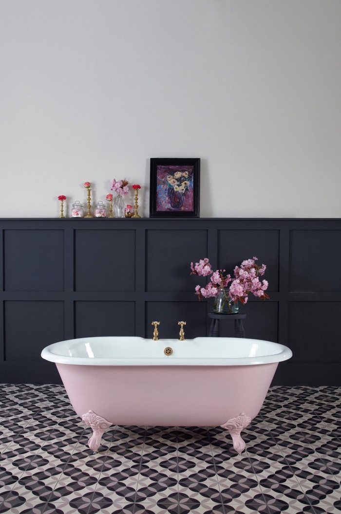 une salle de bains vintage avec un soubassement en panneau mural noir à motifs caissons avec espace rangement en fort contraste avec le mur blanc et la baignoire rose