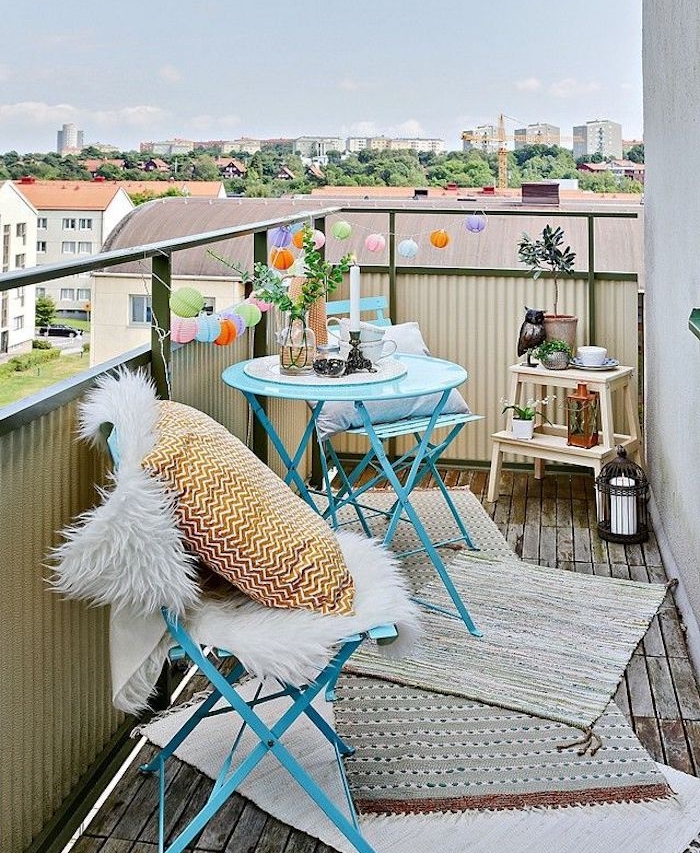guirlande boules colorées sur un balcon aménagé avec table et chaises metal bleues et tapis gris sur sol en bois, petite etagere rangement plantes et deco