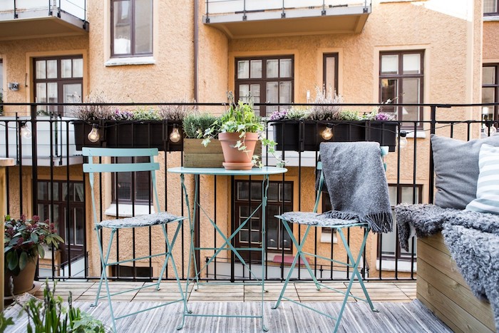 bacs à fleurs suspendus sur le garde corps d un balcon, idée comment aménager son balcon avec chaises et table en metal, tapis gris, banc bois couverte de coussins et plaid moelleux