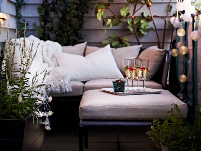 canapé d angle sur une terrasse avec plaid douillet et coussins décoratifs, guirlande boule lumineuse, plantes décoratives, verres à champagne