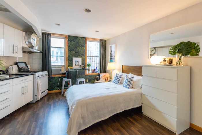 petit appartement une pièce avec lit en face d une cuisine blanche, grande commode blanche, mur d accent décoré de papier peint tropical