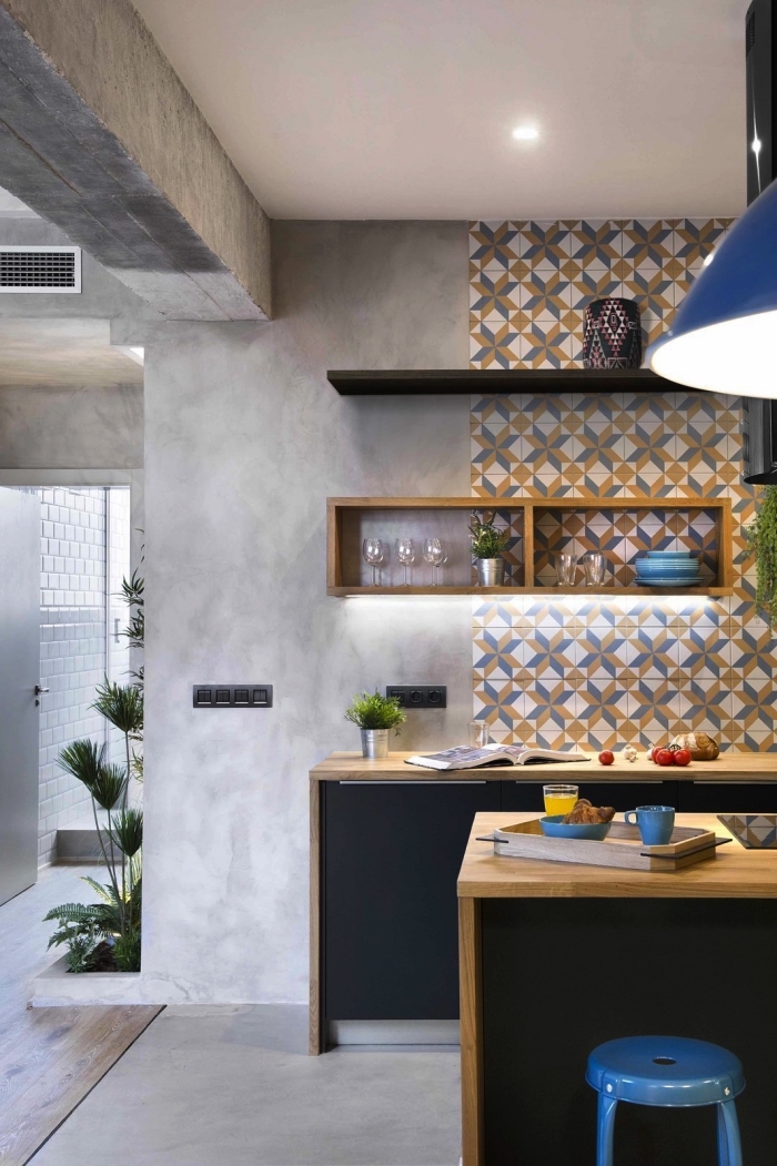 des carreaux de ciment adhesif qui recouvre une partie du mur jusqu'au plafond, en contraste avec les murs effet béton, des meubles de cuisine bois et noir mat