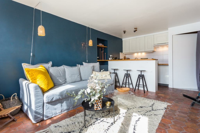 sofa gris, tapis berbère, coussin jaune, peinture murale bleue, lampes suspendues, cuisine petit espace avec îlot, sol en tomettes rouges
