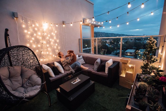 deco hygge de balcon spacieux avec balancelle, cananpé d angle noir avec coussin d assise gris et deco de guirlande lumineuse, revetement balcon d herbe artificielle, plantes decoratives