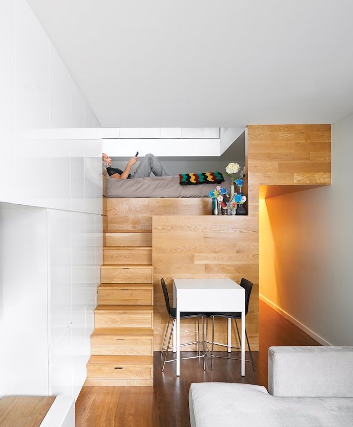 design minimaliste dans un studio tout petit en bois avec lit en dessus d une cuisine, table blanche et chaises noires, canapé gris clair