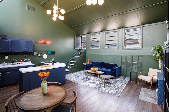 murs vert olive, cuisine, ilot central et canapé bleu foncé, parquet marron foncé, table bois ronde entourée de chaises anciennes, tapis gris et blanc