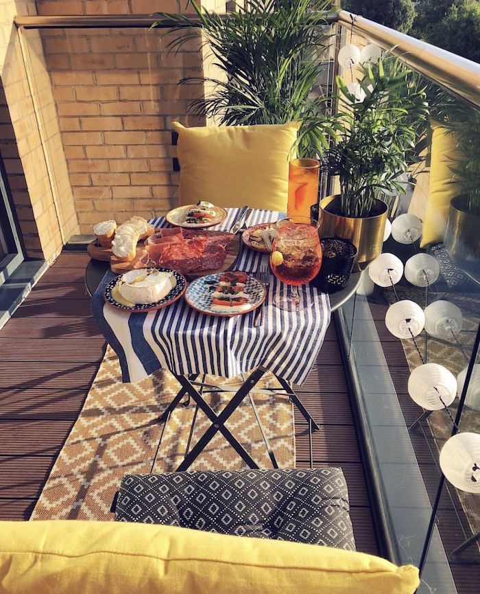 petit dejeuner servir sur le balcon, chaises rangées autour d une table ronde, tapis jaune, garde corps en verre, guirlande boule, plantes exotiques