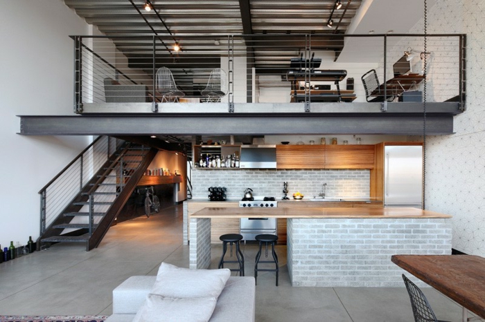 mezzanine avec rambarde noire, escalier métallique, ilot en bois sur muret gris, tabourets industriels, coin travail et chambre à coucher