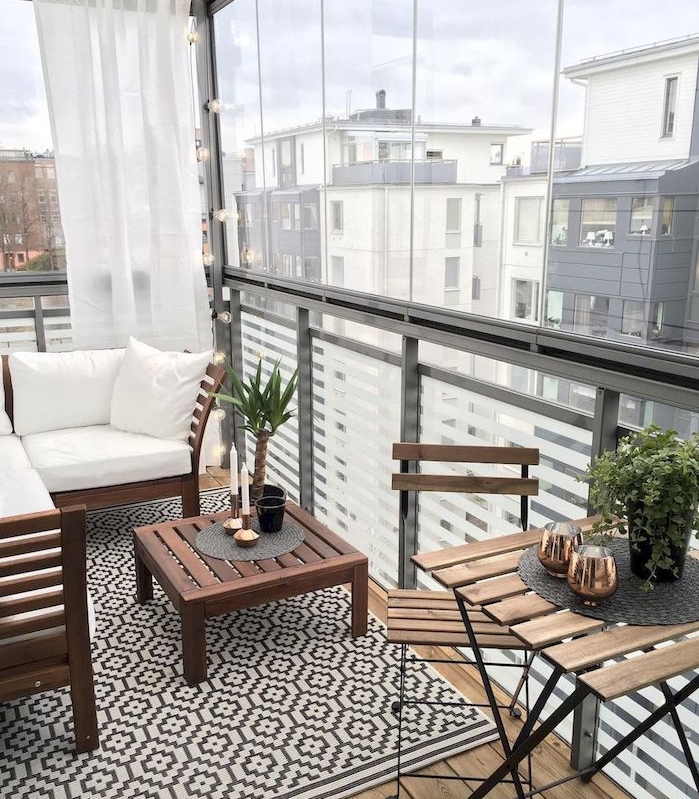 petit coin repos en canapé et table bois sur un balcon vitré avec brise vue rideau blanc, tapis gris et blanc et table et chaises bois et metal sur baclon bois