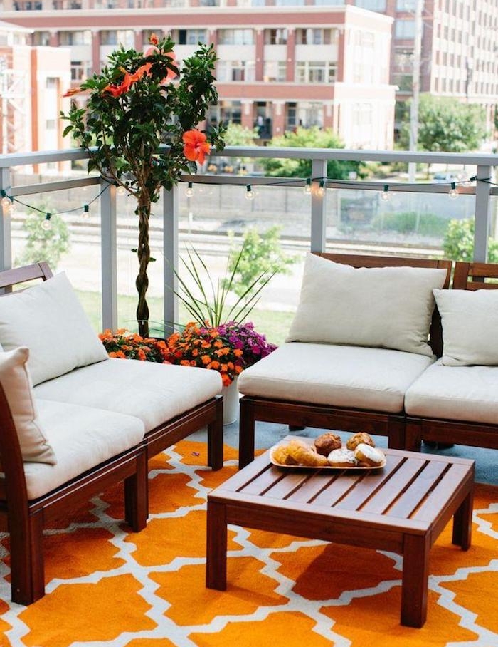 salon de balcon coloré avec canapés en bois et table bois sur tapis orange, fleurs en pots et guirlande lumineuse originale