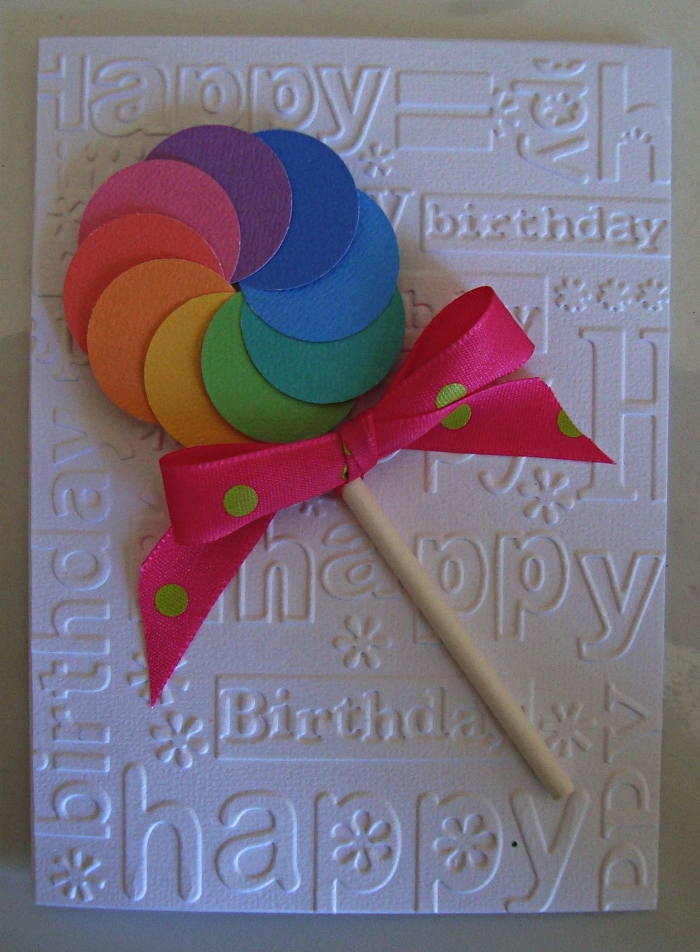 carte anniversaire 1 an en papier blanc embossé décorée d'une sucette en papier cartonné aux couleurs de l'arc-en-ciel