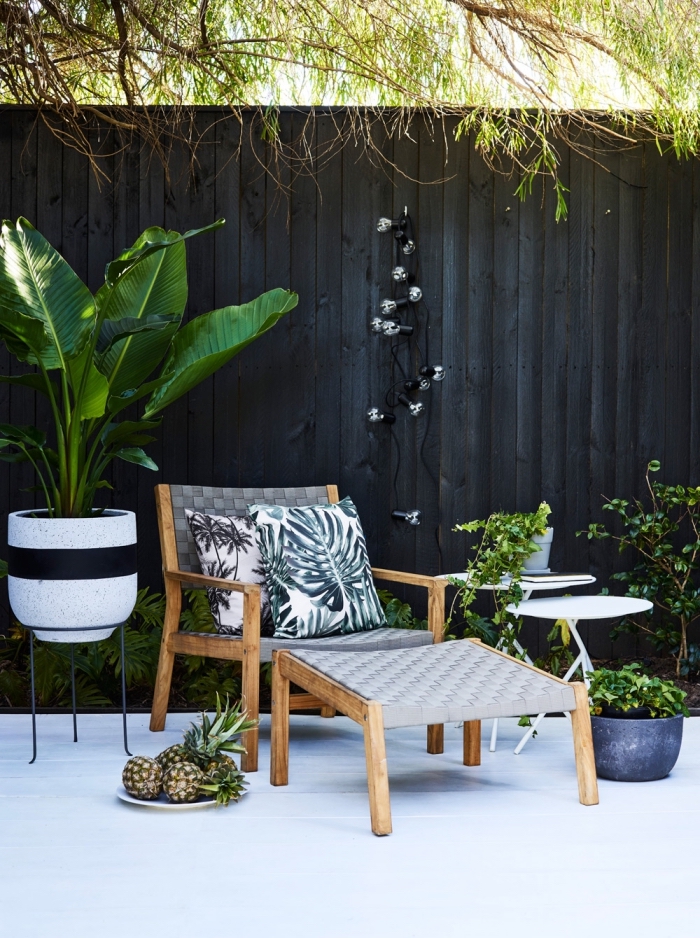 idee amenagement jardin avec palissade repeinte en noir et terrasse blanche, déco tropicale avec plantes exotique et coussins à motifs feuilles