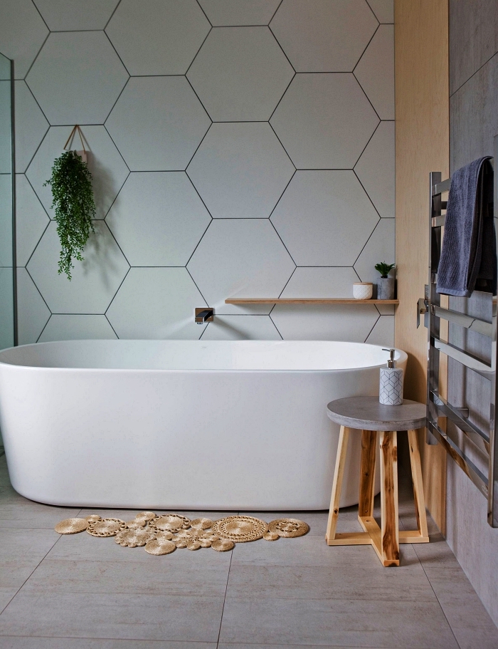 une salle de bain blanche avec baignoire ovale moderne contre un mur en panneau mural à motifs hexagonaux, déco naturelle avec tapis bohème en fibres végétales et table d'appointe en bois et béton