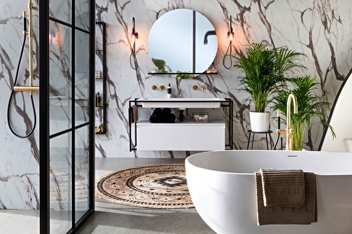 salle de bain contemporaine en marbre avec baignoire îlot aux lignes modernes et douche italienne délimitée par une verrière industrielle, salle de bains moderne esprit minimaliste et nature décorée de plantes vertes et d'un tapis rond en fibres naturelles