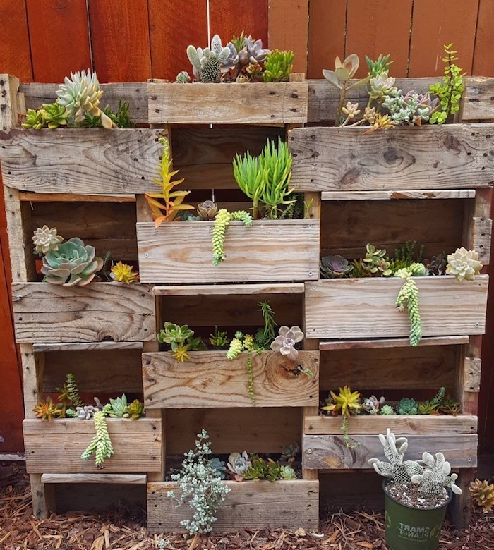 plusieurs bacs à fleurs en bois assemblés dans un rangement plantes exterieur adossé à un mur de cloture en bois