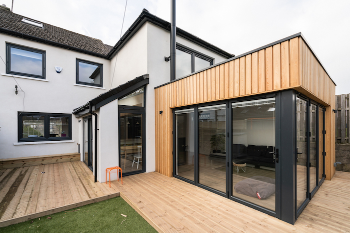 agrandissement maison bois avec à baies vitrées avec canapé noir et table basse, installation extension sur terrasse exterieure de teck