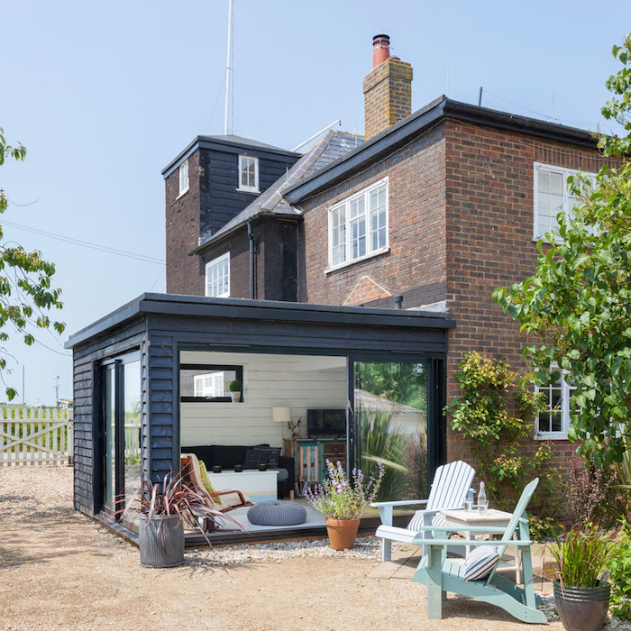 modele de maison de briques avec une veranda adossée en alu couleur noire, amenagement salon avec chaise longue, table basse et canapé gris sur véranda