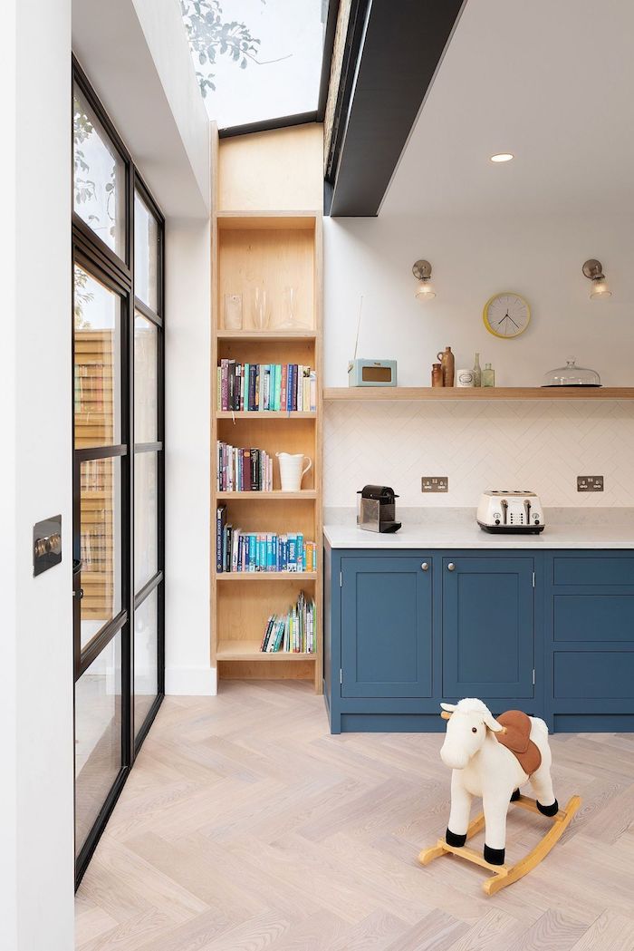 cuisine bleu foncé et bibliotheque d angle à coté d une verrière extérieure industrielle, parquet bois clair, murs blancs, étagère de bois