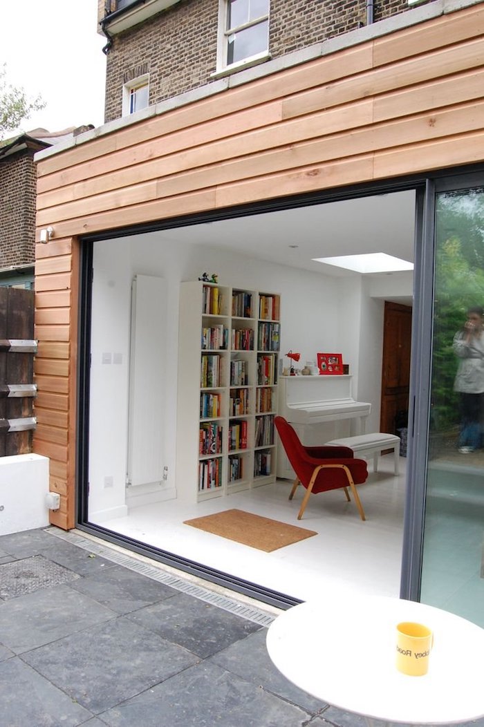 coin salon minimaliste avec bibliotheque scandinave blanche, chaise rouge et piano blanc accueillis par une extension en bois à vitraux